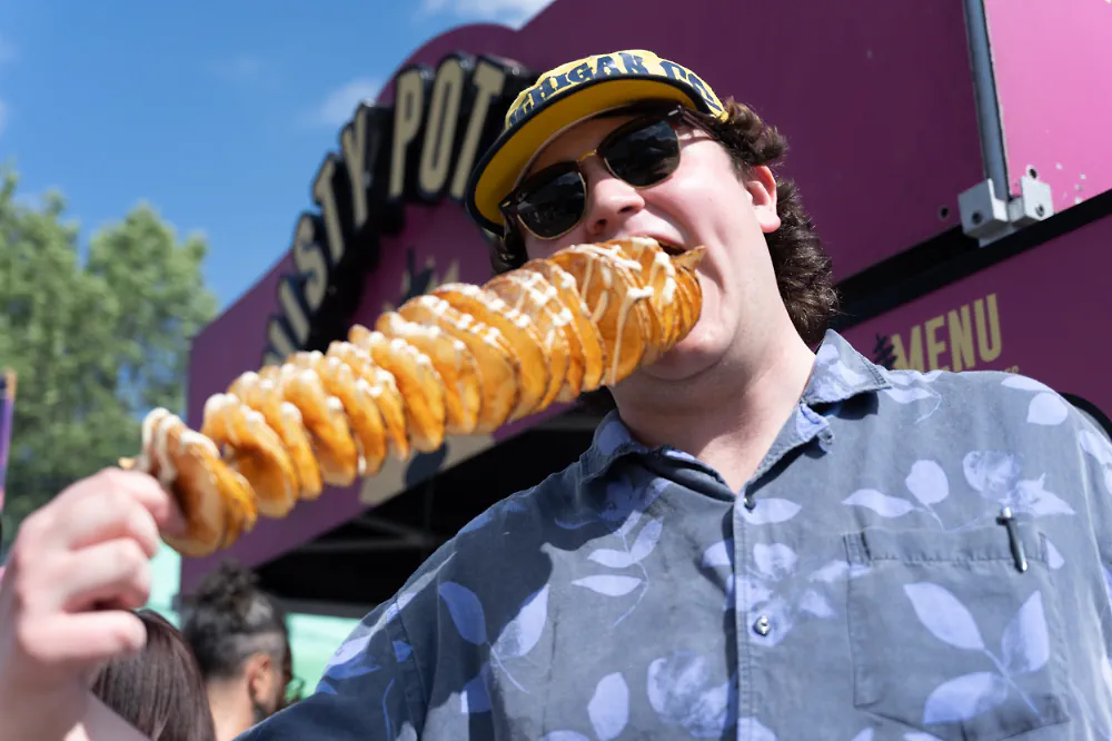 Festivalgoer biting a giant spiral potato on a stick at a food vendor stall.